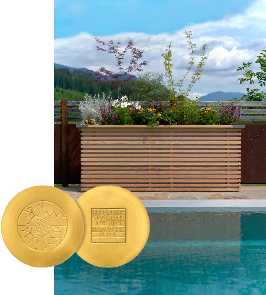 Modernes Edelstahl Pflanzengefäß mit Holzverkleidung, bepflanzt mit Blumen, vor Berglandschaft und Pool. Im Vordergrund zwei goldene Medaillen des Bayerischen Staatspreises für das Handwerk